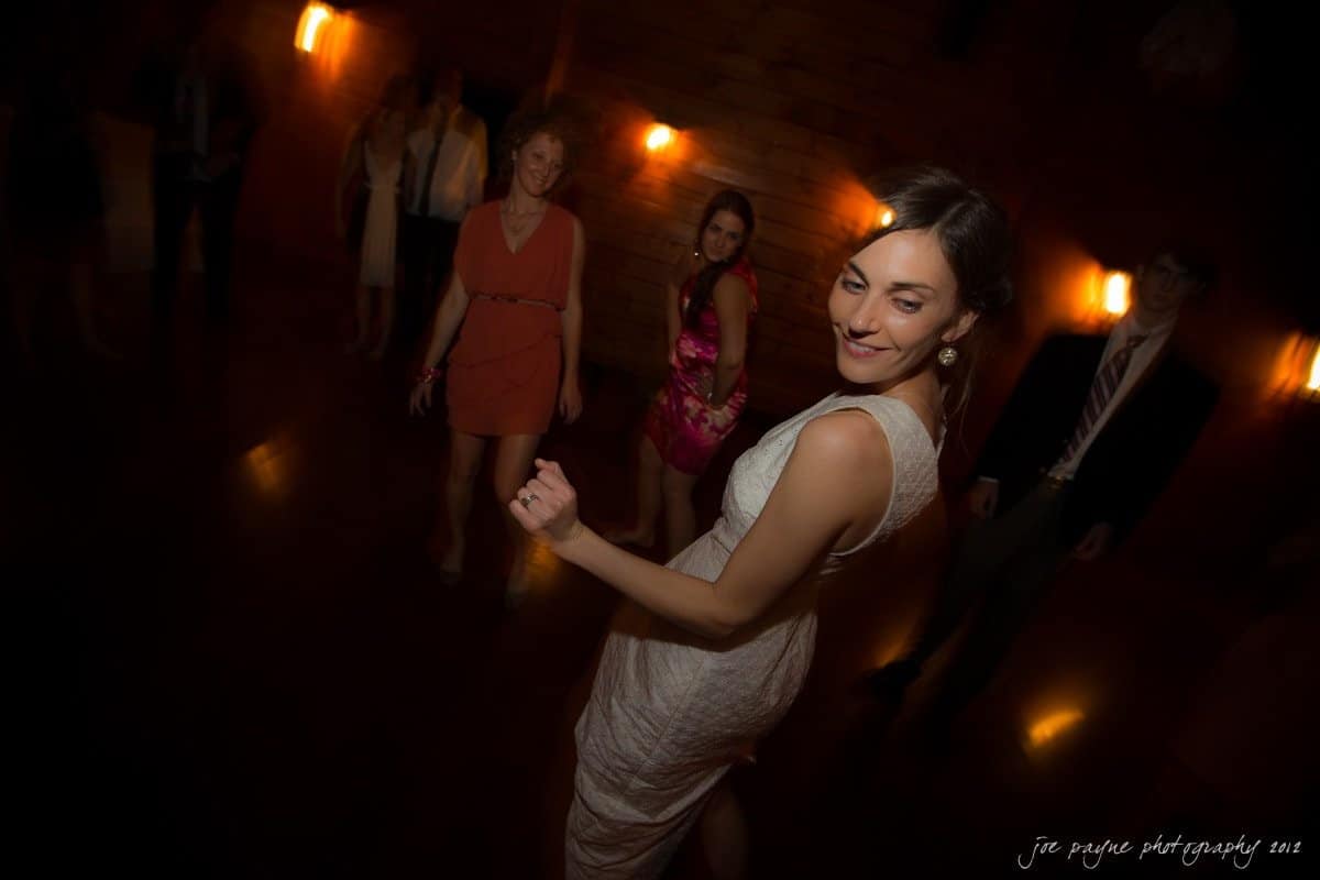 barn at valhalla, chapel hill wedding photography ~ allegra and chris A woman in a white dress smiles as she dances in a dimly lit room. Other people in colorful dresses and suits dance in the background, illuminated by soft, warm lighting. The atmosphere appears to be festive and lively.