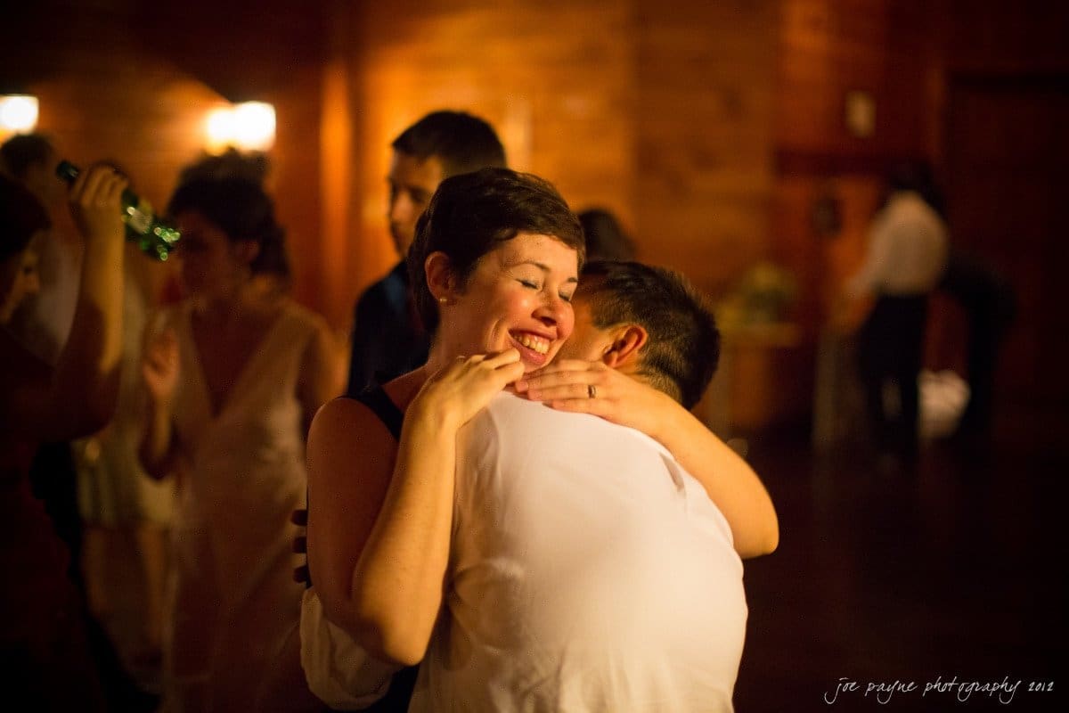 barn at valhalla, chapel hill wedding photography ~ allegra and chris A joyful woman with short hair embraces a man in a white shirt on a dance floor. The background is warmly lit, with other people dancing and lights softly glowing in the dim room.