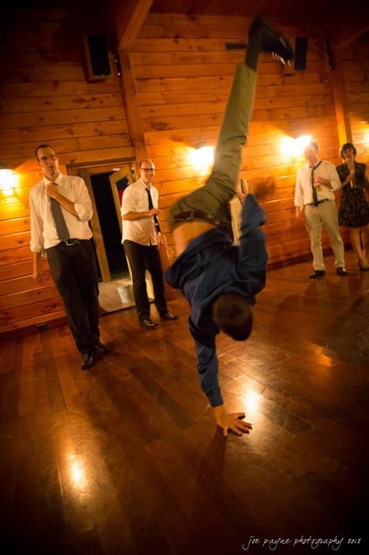 barn at valhalla, chapel hill wedding photography ~ allegra and chris A person performs a one-handed handstand while others watch in a wooden hall with dim lighting. Several people are dressed in formal attire.