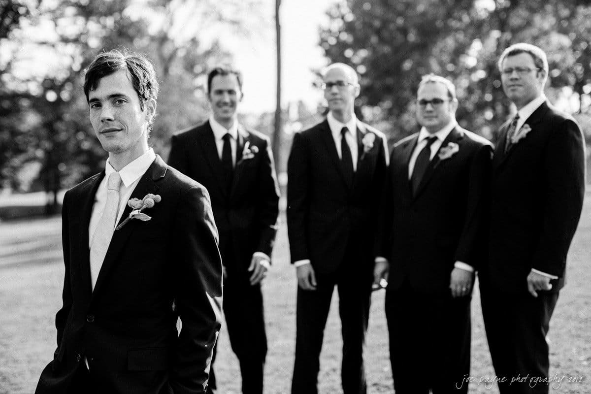 barn at valhalla, chapel hill wedding photography ~ allegra and chris A groom stands in the foreground, looking at the camera, while four groomsmen in suits stand behind him, slightly out of focus. They are outdoors on a sunny day with trees in the background. Black and white image.