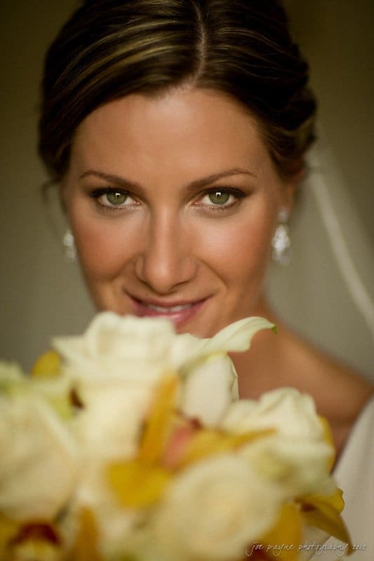 A bride with light brown hair and sparkling eyes smiles gently, holding a bouquet of white and yellow flowers close to her face. Her earrings and veil are visible, adding to her elegant appearance.