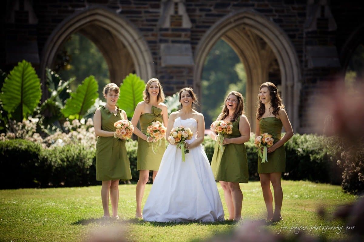 A bride in a white dress stands outdoors with four bridesmaids in green dresses, all holding bouquets. They are in front of a stone structure with arched openings and surrounded by greenery. The sun is shining brightly.