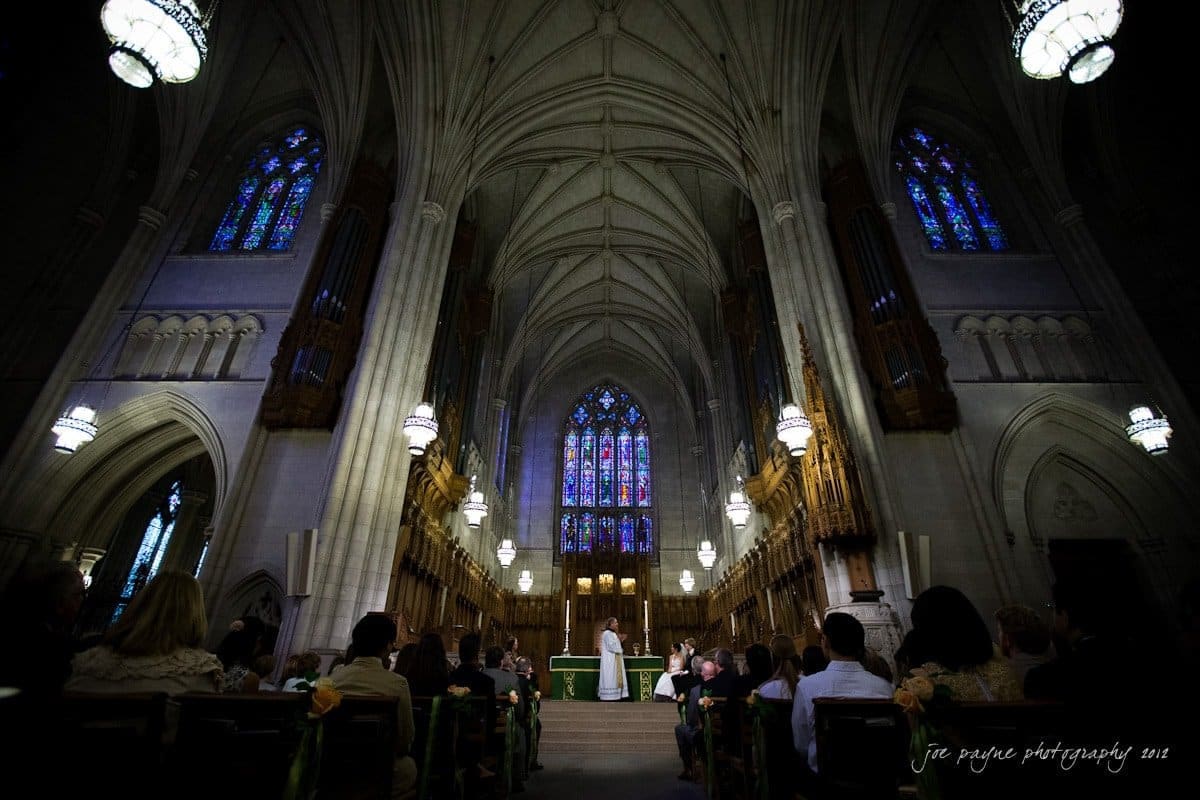 A couple stands at the altar in a grand cathedral with high vaulted ceilings and stained glass windows, surrounded by seated guests.