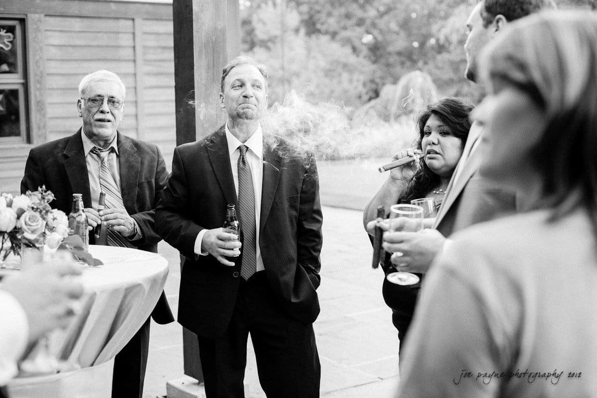 A black and white photo of several people standing outdoors in formal attire, holding drinks and smoking. A table with a flower arrangement is on the left. The scene appears relaxed and social.