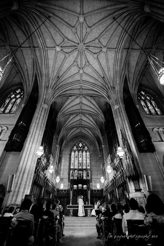 A black and white photograph of an ornate cathedral interior with a high, vaulted ceiling. A couple stands at the altar during a wedding ceremony, surrounded by seated guests. Stained glass windows are visible in the background.