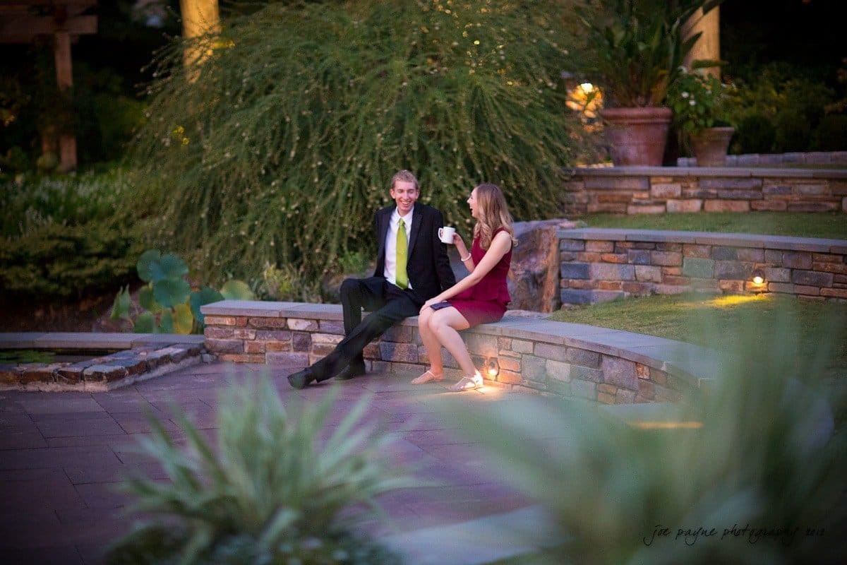 A man and a woman sit on a stone ledge in a garden, engaging in conversation. The woman holds a cup. The scene is softly lit with decorative garden lights, surrounded by greenery and stone walls.