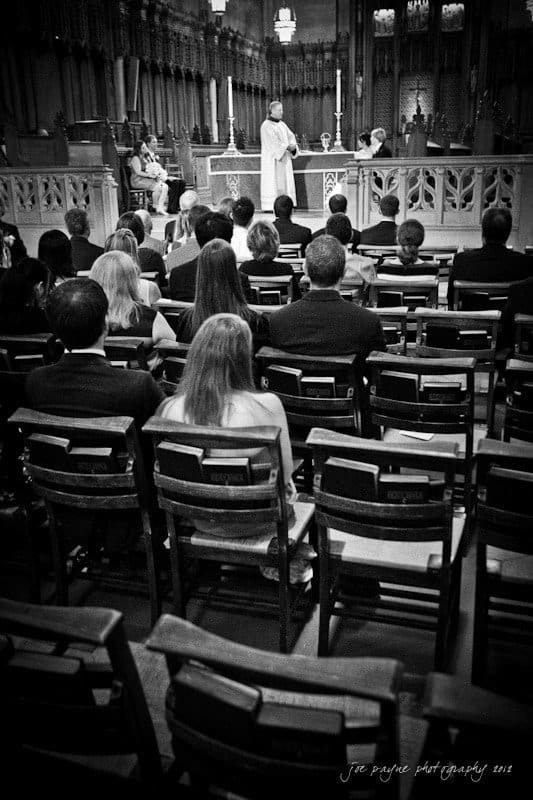 A black and white photo of a wedding ceremony in a church. The bride, groom, and guests are seated as a clergyman stands at the altar. The church features ornate wooden details and candles.