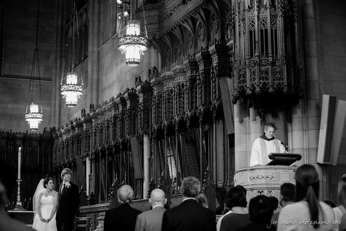 A black and white photo of a wedding ceremony in a grand church. A bride and groom stand before an altar, facing a minister who is speaking. The church features intricate Gothic architecture and ornate hanging lights, with guests seated in the foreground.