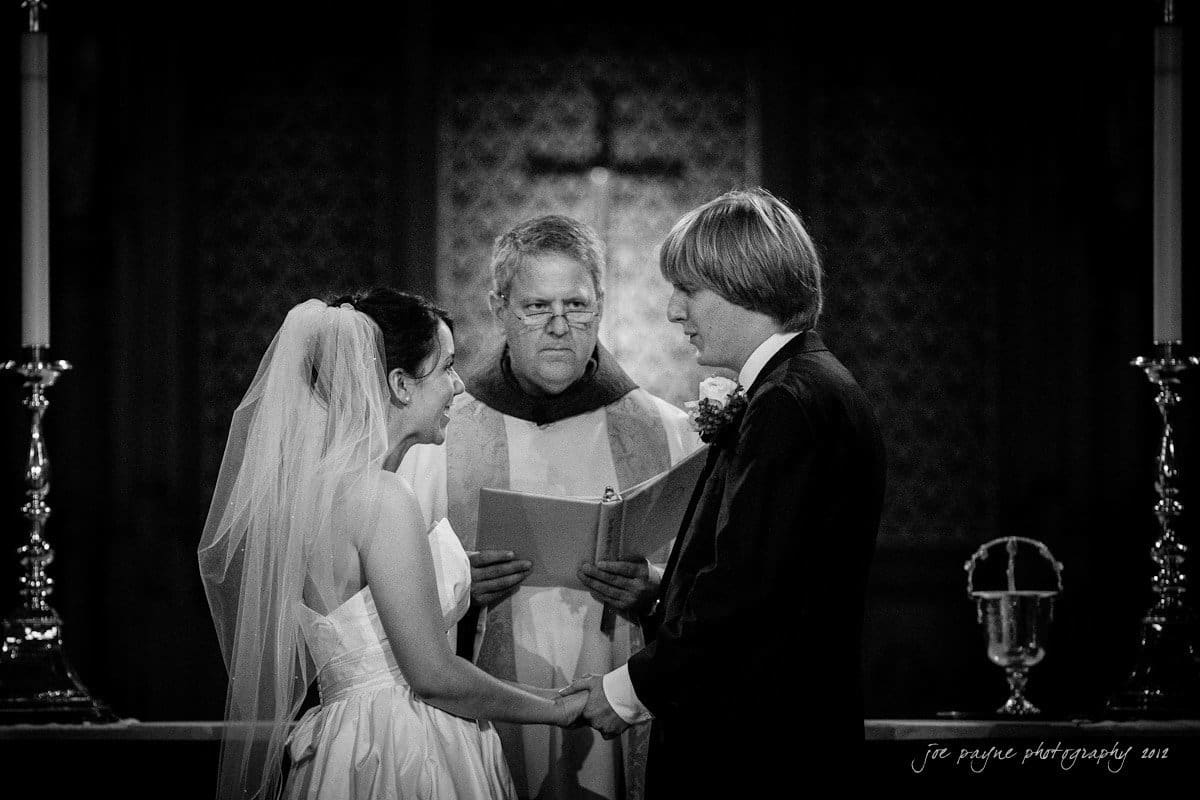 A bride and groom stand facing each other, holding hands during a wedding ceremony. The officiant, holding a book, looks on. The setting is a dimly lit church altar, with candles on either side.