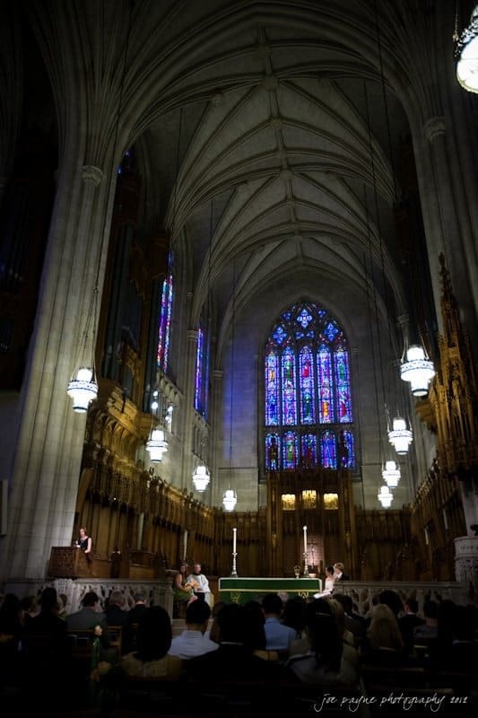 A grand cathedral interior with a high vaulted ceiling and colorful stained glass windows. Ornate chandeliers hang from the ceiling, and people are seated below, facing a central altar. The atmosphere is serene and reverent.