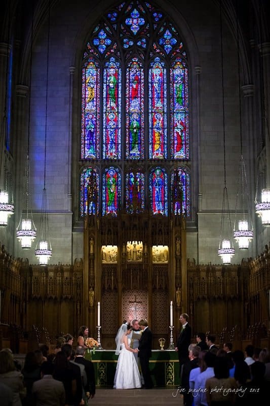 A couple exchanges vows in a grand church with a large, vibrant stained glass window as the backdrop. The ornate wooden interior is softly illuminated. Guests are seated, witnessing the ceremony.