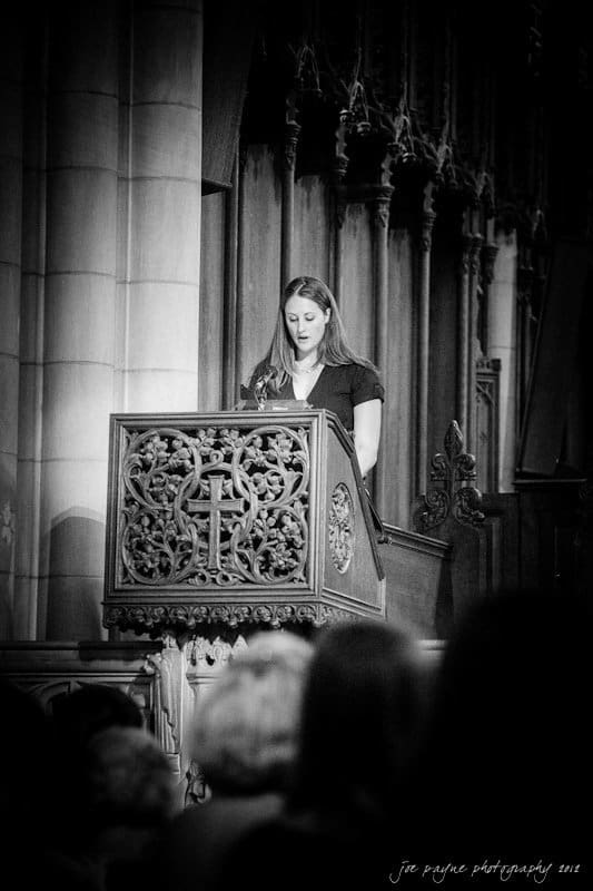 A woman stands at a carved wooden lectern with a cross design, reading or speaking in a dimly lit, ornate church interior. The background features tall, detailed pillars and arches.