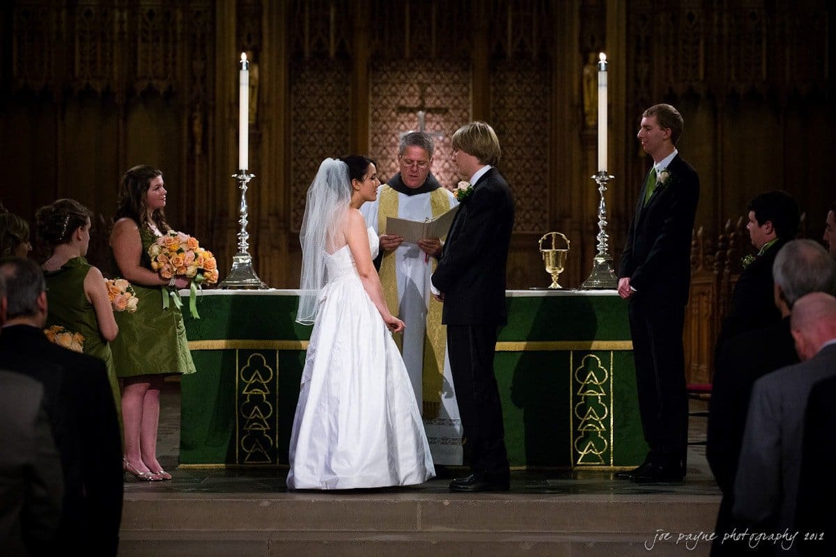 A bride and groom stand at the altar during their wedding ceremony in a church. Bridesmaids in green dresses and groomsmen in dark suits with green ties stand to the sides. A priest officiates, holding a book. Candles and an ornate backdrop are visible.