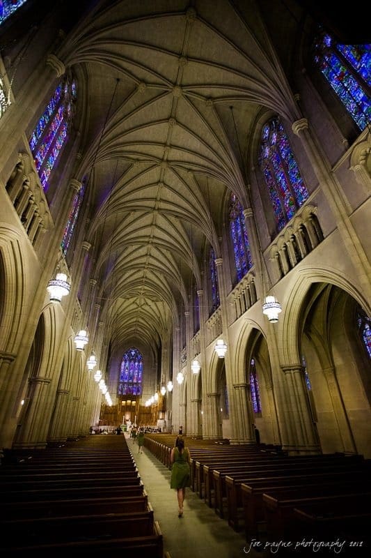 A person walks down the aisle of a grand church interior with vaulted ceilings, ornate stained glass windows, and hanging chandeliers. The architecture features Gothic elements, and the atmosphere is serene and majestic.