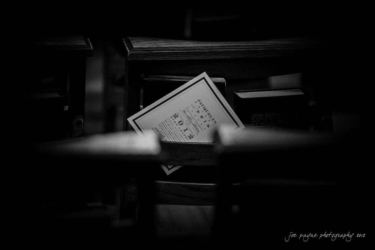 A black and white image of a church pew with hymnals and a folded bulletin placed within. The pew is dark, with a focus on the papers, creating a contemplative atmosphere.