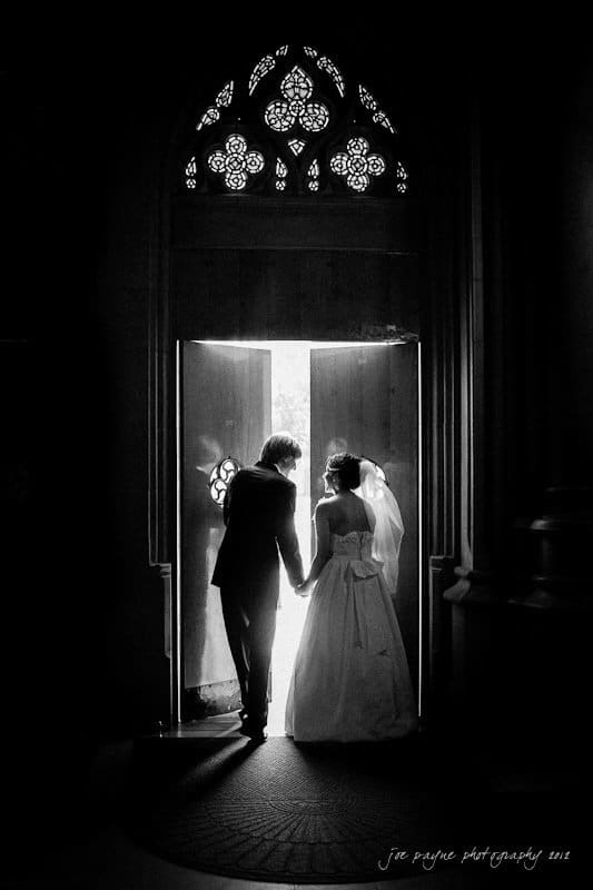 A bride and groom stand hand in hand, backlit by light streaming through an open door in a dark room. The bride wears a long gown and veil, and the setting has ornate architectural details.