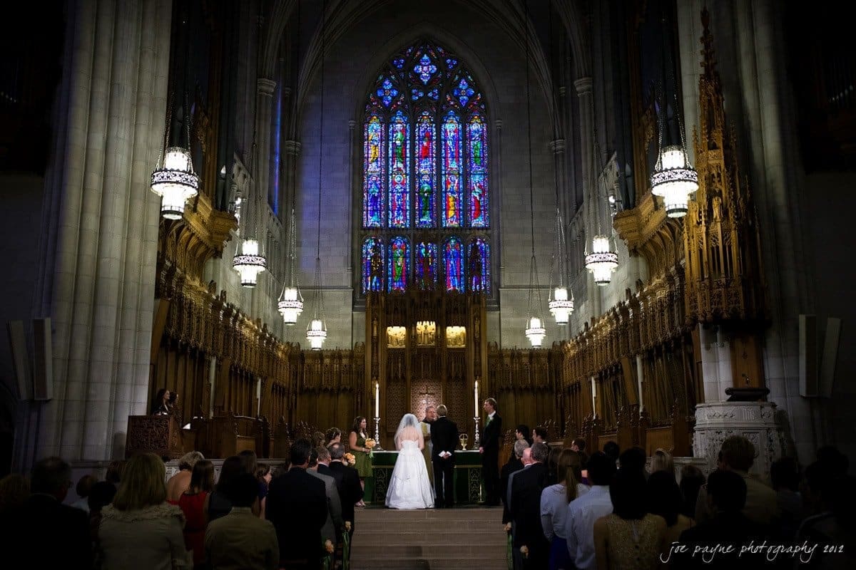 A wedding ceremony takes place inside a grand cathedral featuring gothic architecture. The bride and groom stand before an altar, illuminated by vibrant stained glass windows behind them. Guests seated on both sides witness the event.
