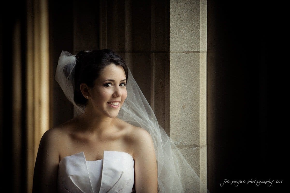 A bride in a white dress stands by a window, smiling softly. Her veil is draped over her shoulder, and soft light highlights her features against the textured stone background.