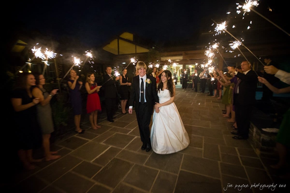A bride and groom walk down a stone path at night, smiling, surrounded by guests holding sparklers. The bride wears a white gown and the groom is in a black suit. The scene is festive and joyful, with warm lighting.