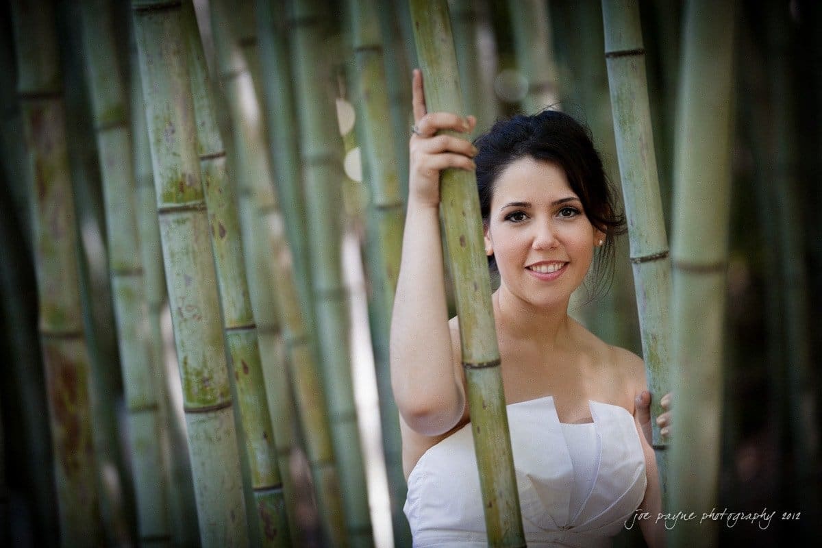 A woman in a white dress stands among tall bamboo stalks, smiling warmly at the camera. The soft lighting creates a serene and natural atmosphere.