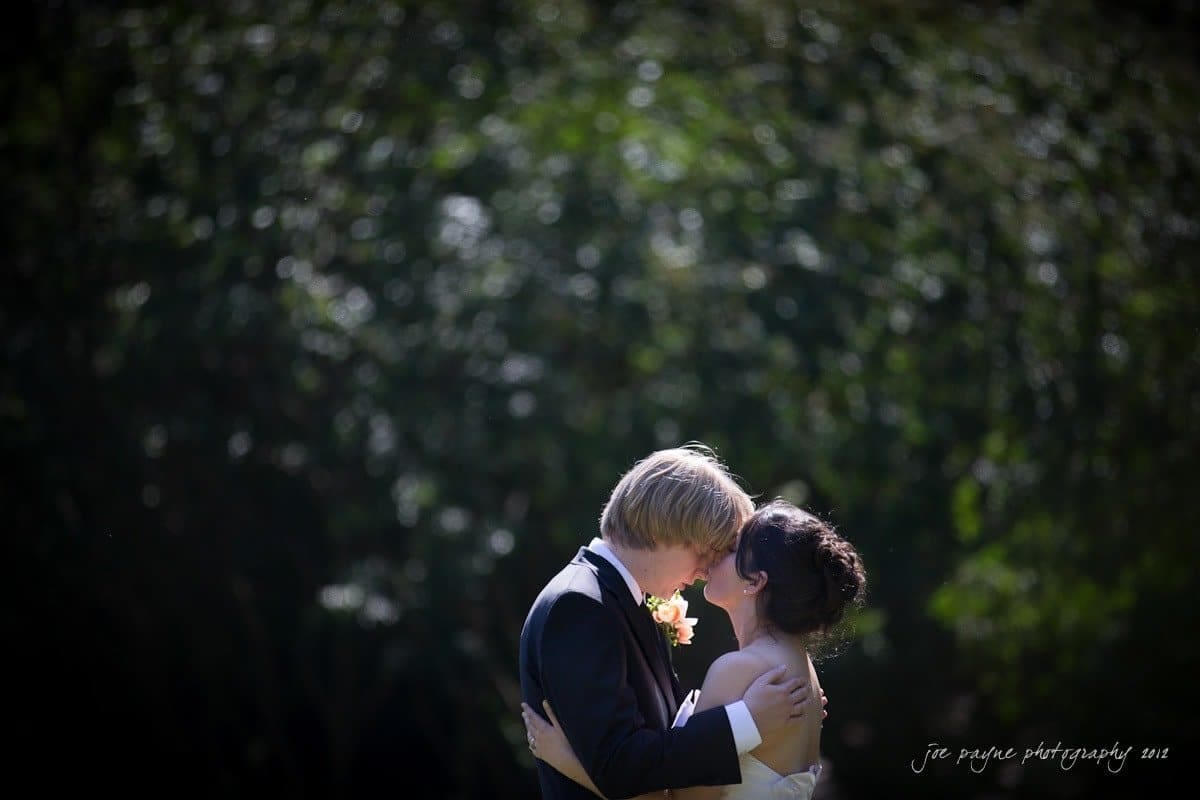 A couple in formal attire embraces outdoors with a lush, green backdrop. The man wears a black suit with a light pink boutonniere, while the woman is in a sleeveless white dress. Their heads touch gently, creating a serene and intimate moment.