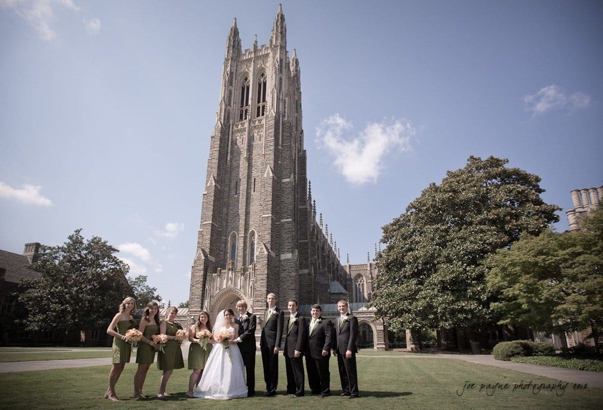 A bride and groom stand with their bridal party in front of a large cathedral with a tall tower. The bridesmaids wear green dresses, and the groomsmen wear black suits. The sky is clear and blue, and the setting is a grassy, open area.