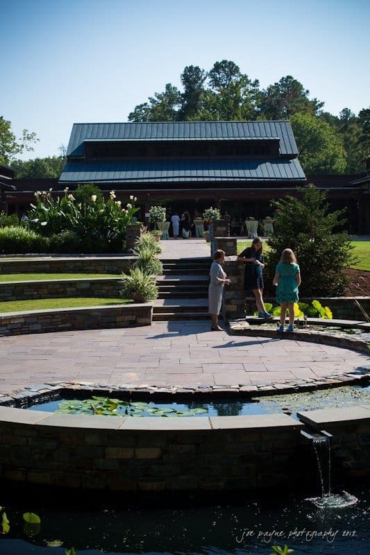 A serene outdoor scene with a group of people gathered near a pond surrounded by lush greenery. A stone pathway leads to a building with a dark roof, set against a backdrop of trees under a clear sky.