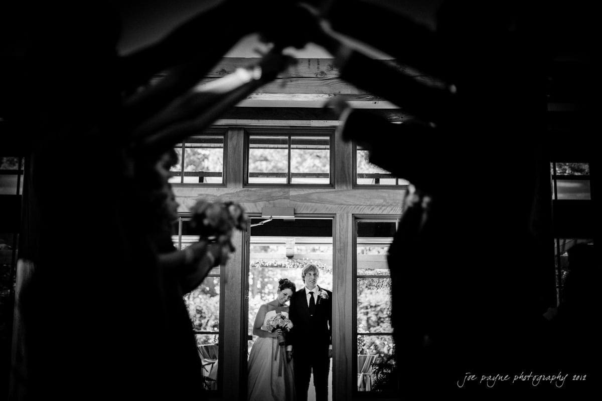 Bride and groom standing together in the background, framed by the arms and hands of people forming an arch in the foreground at a wedding. The style is black and white.