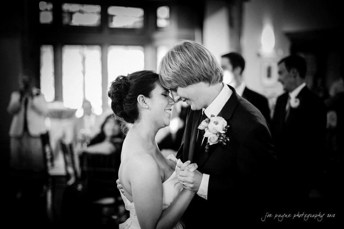 A happy couple dancing closely, smiling at each other during their wedding reception. The bride wears a strapless gown, and the groom is in a suit with a boutonniere. The background shows guests and a warmly lit room.