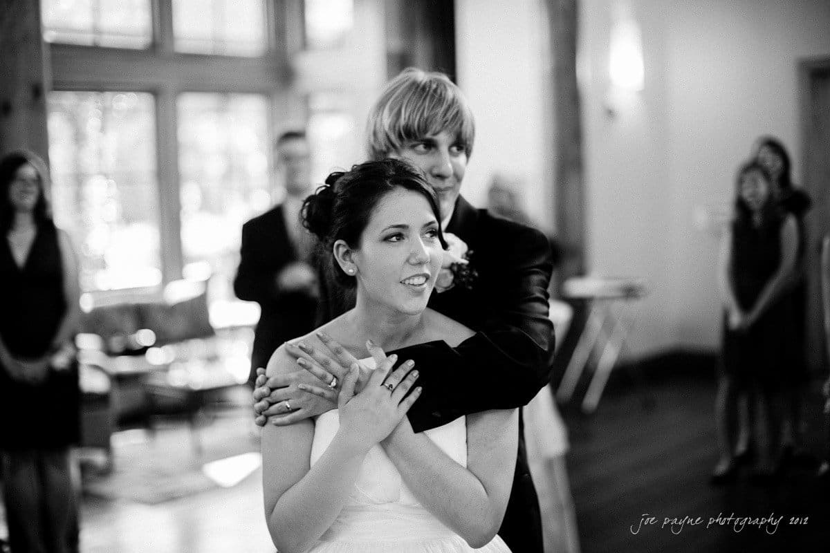 A black-and-white photo of a wedding reception shows a couple embracing. The woman wears a wedding dress and smiles while the man hugs her from behind, wearing a suit. Blurred guests are visible in the background.