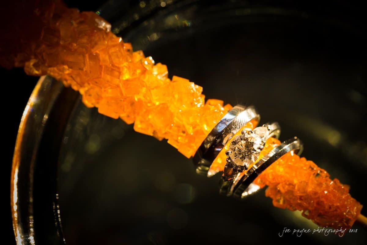 Close-up of a diamond ring resting on bright orange sugar crystals. The silver band contrasts with the vibrant sugar, creating a striking visual effect. The background is softly blurred.