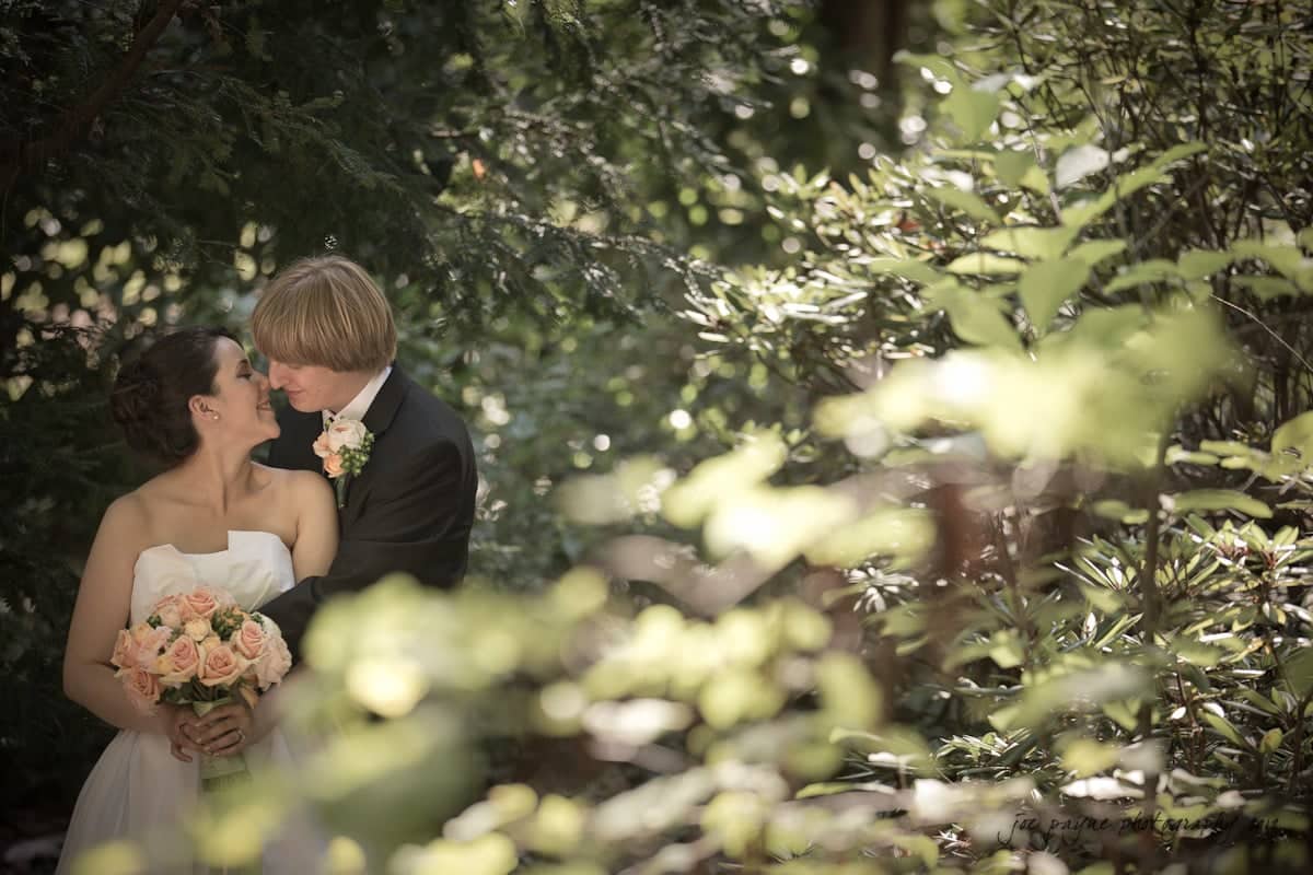 A bride and groom share a tender moment in a lush, green forest setting. The bride holds a bouquet of pink and peach roses, and dappled sunlight filters through the trees, creating a serene atmosphere.