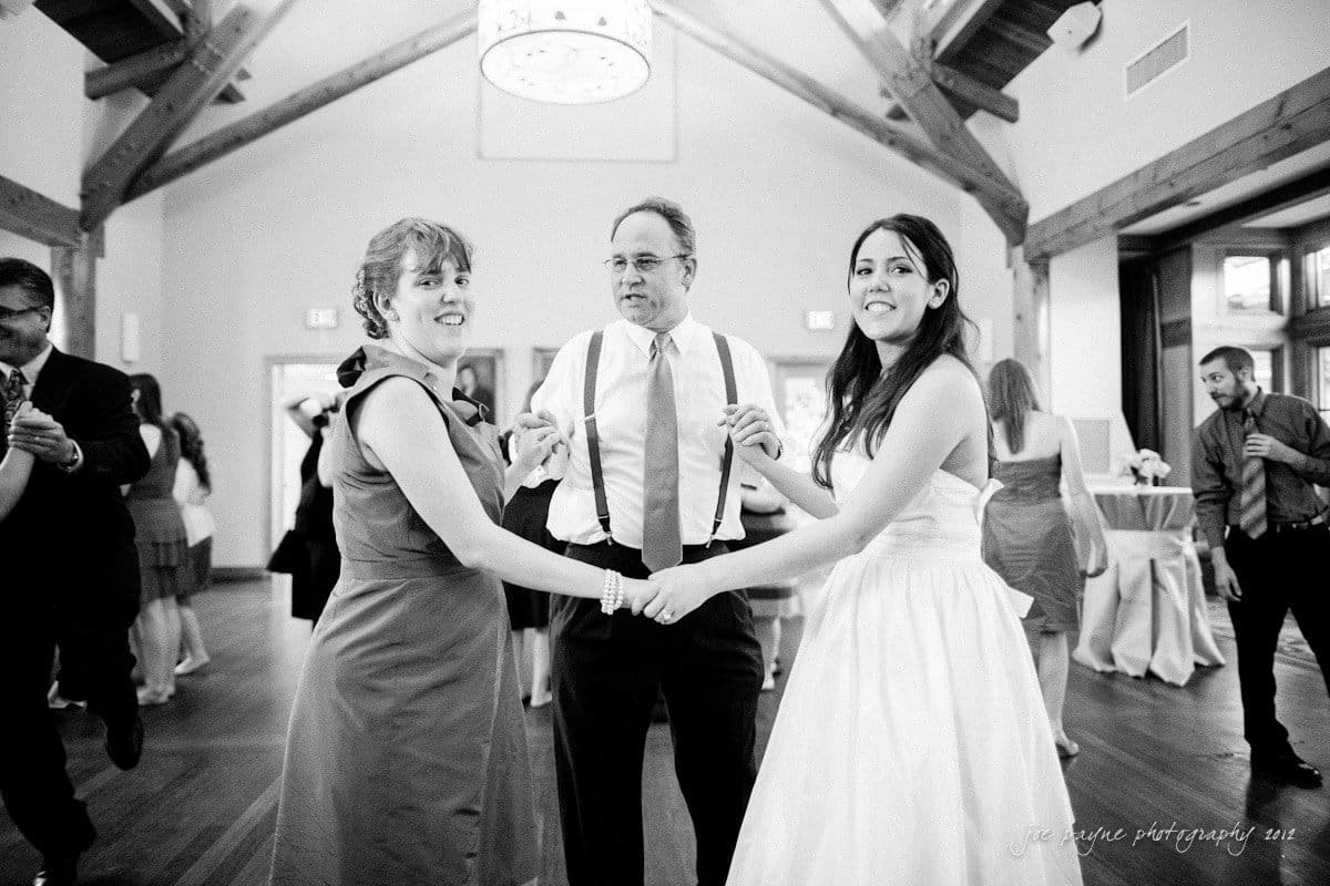 A bride in a white dress and two guests, a woman in a dark dress and a man in a shirt with suspenders, hold hands in a circle while dancing at a wedding reception. The setting is indoors with wooden beams.