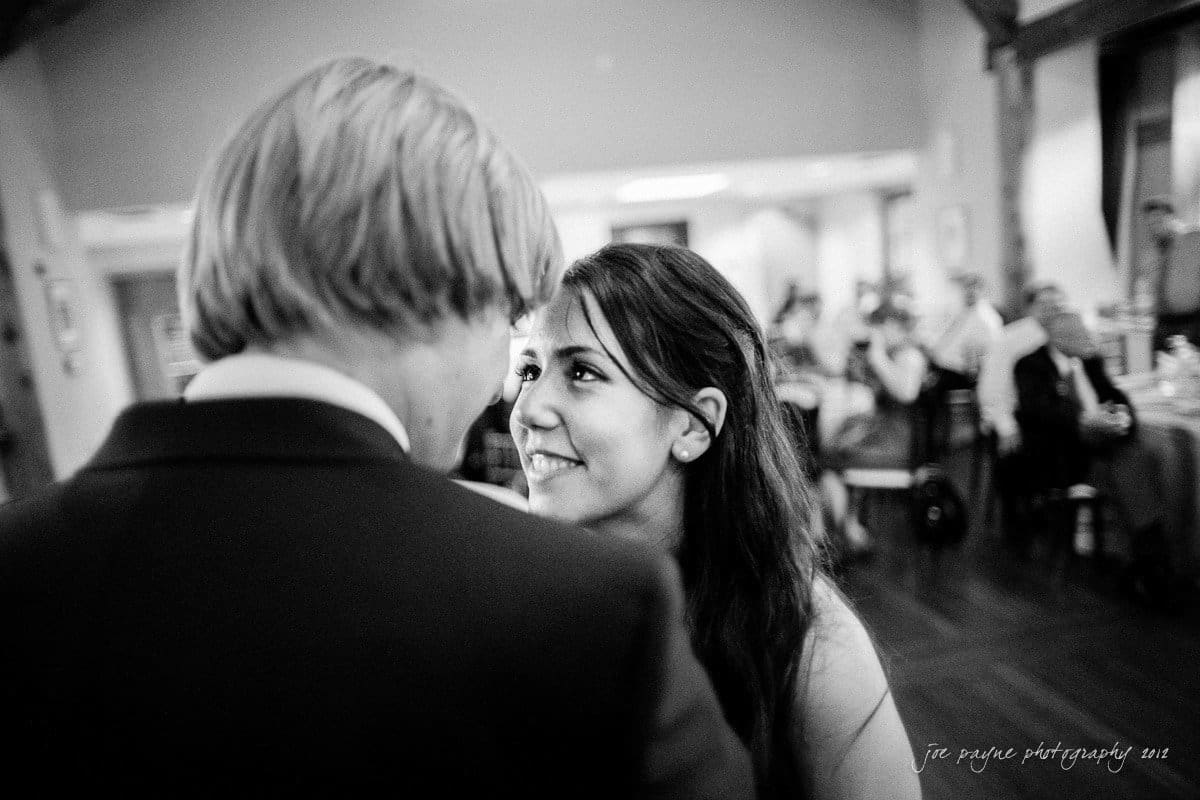 A black and white photo of a woman smiling at a man with blond hair in a dimly lit room. People are seated in the background, creating a warm, intimate atmosphere.