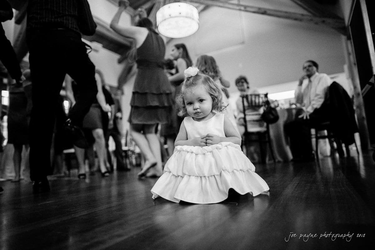 A young girl in a white dress crouches on a dance floor while adults dance around her. The background is blurred, highlighting the movement and dance scene in a lively indoor setting.