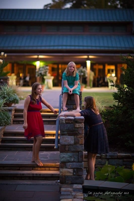 Three young girls are interacting on a stone staircase in the evening. One girl sits atop the railing, while the other two stand chatting. The background features a building with a green roof and warmly lit windows.