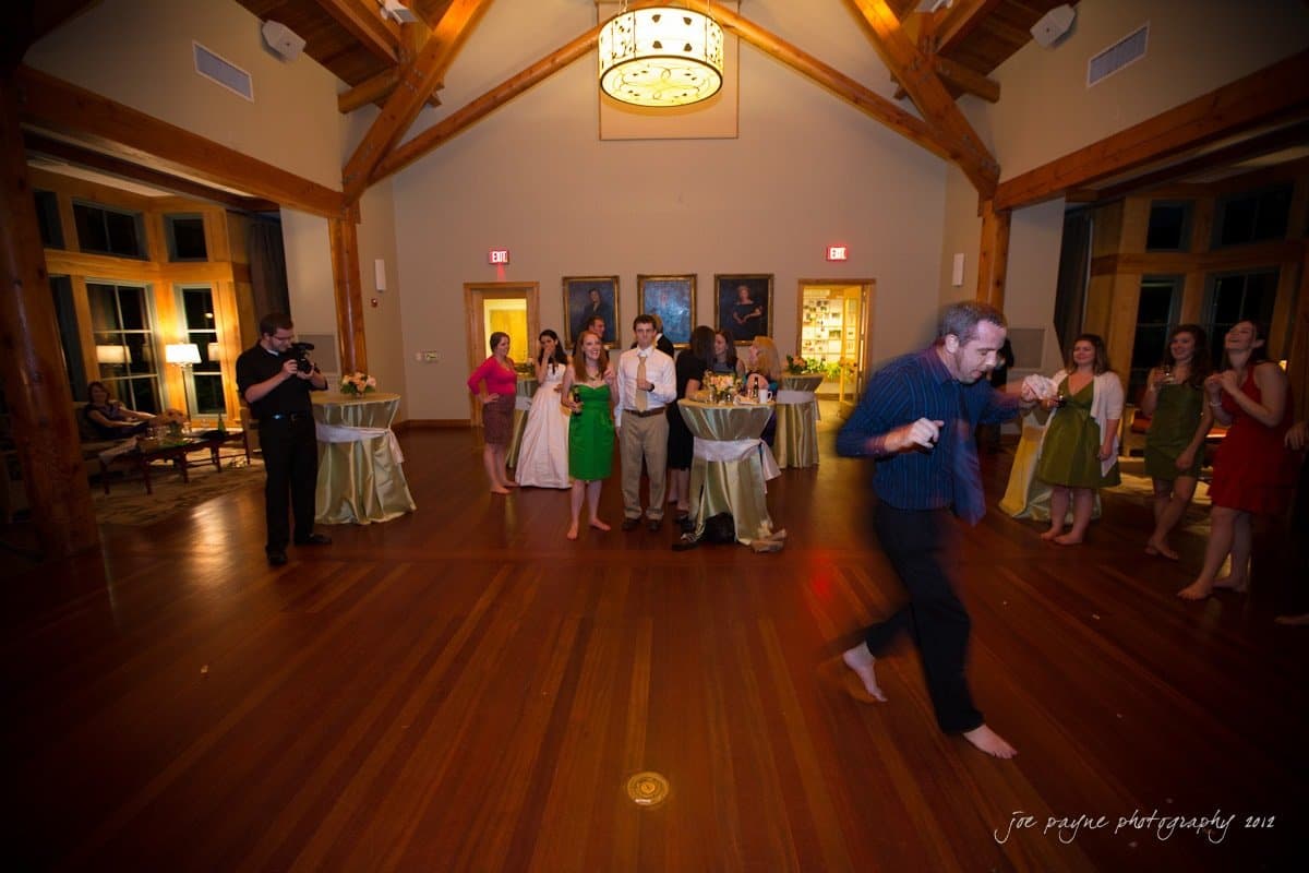 A person dances barefoot in the center of a warmly lit room with wooden floors. Guests stand around, watching and chatting near tables with white tablecloths and flower arrangements, under a ceiling with wooden beams.