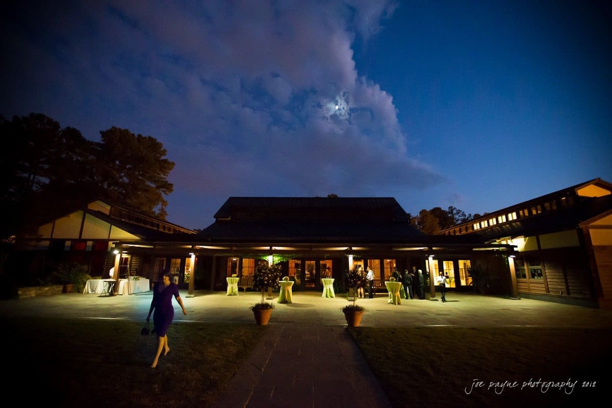 A person in a blue dress walks towards a large building at night, with glowing lights visible through the windows. Several cocktail tables are set up outside. The sky is partly cloudy with the moon peeking through.