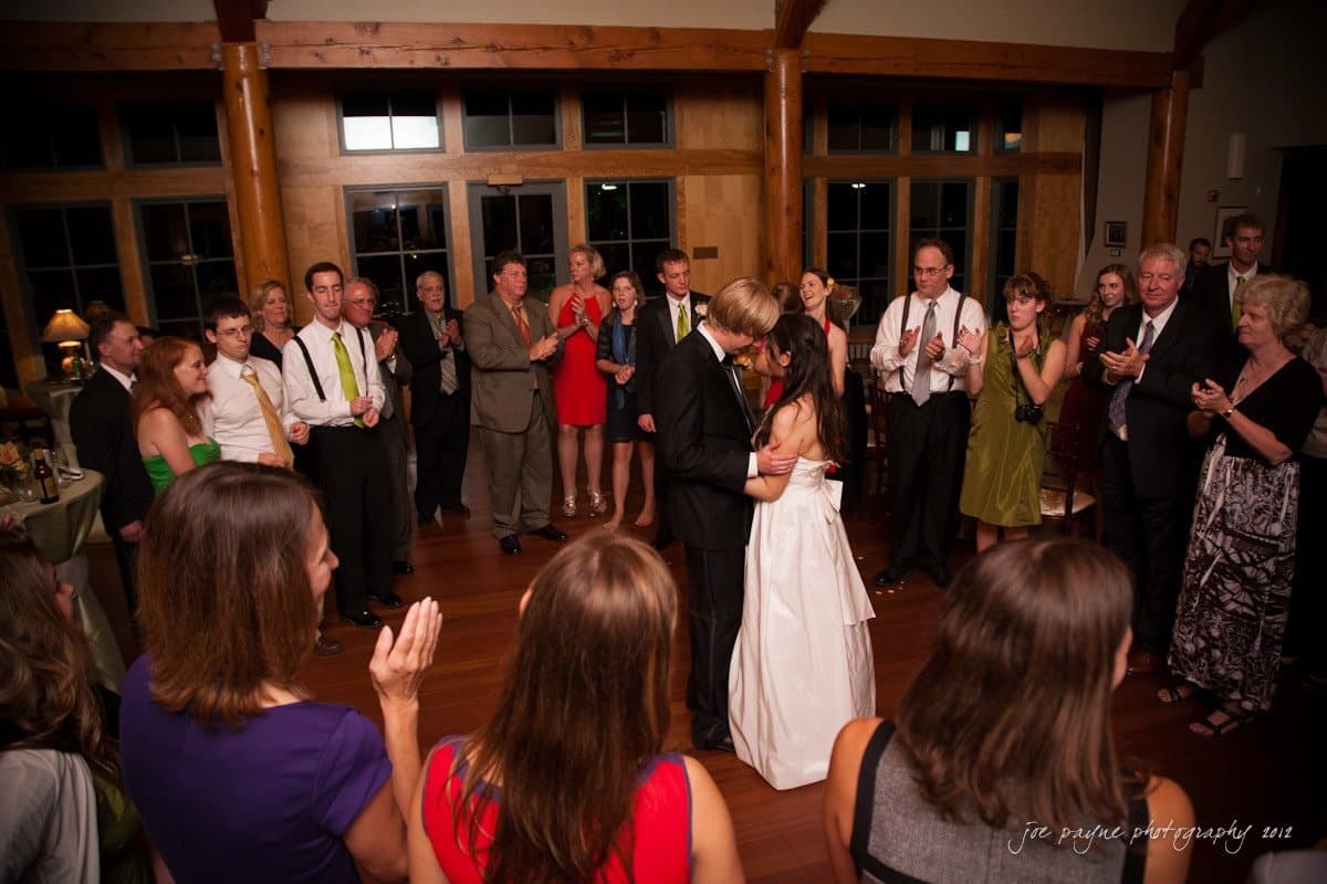 A newlywed couple shares their first dance at a wedding reception. They are surrounded by guests who are clapping and watching them. The venue features wooden beams and large windows, creating a warm atmosphere. The bride wears a white dress, and the groom is in formal attire.