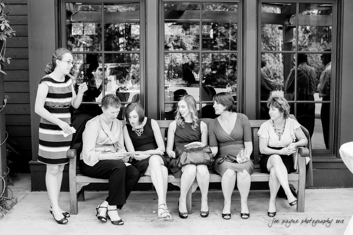 A group of six women, dressed formally, are gathered around a bench. Five are seated, engaged in conversation or observing a device. One stands nearby, smiling. They are in an outdoor setting, with glass windows reflecting trees in the background.