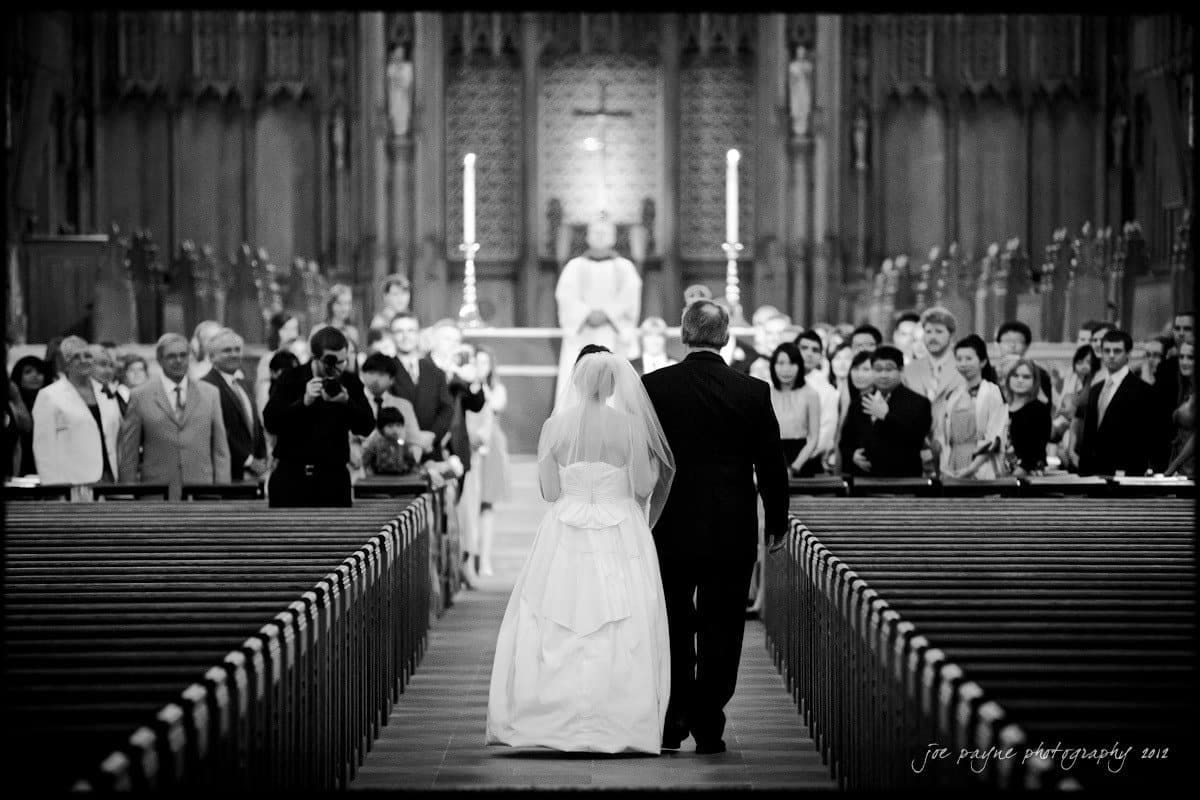 A bride in a white dress and veil walks arm in arm with a man in a suit down the aisle of a large, ornate church. Guests stand on either side, watching the procession, with a priest and altar visible ahead.