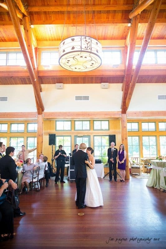 A bride and groom share their first dance in a wooden-beamed reception hall with large windows. Guests sit and stand around watching, and a photographer captures the moment. A large chandelier hangs overhead.