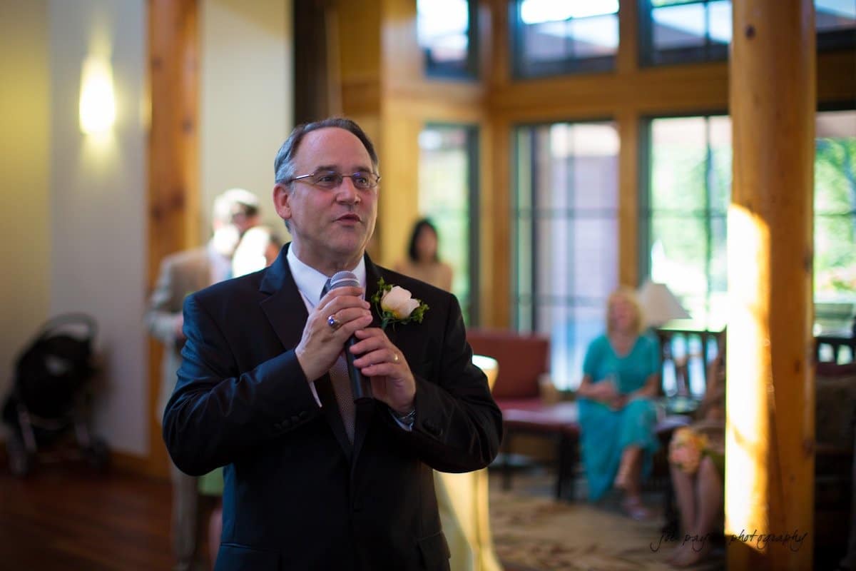 A man in a suit and tie holds a microphone, speaking at an indoor event. He has a flower boutonniere on his jacket. The room has large windows and wooden pillars, with people seated in the background.