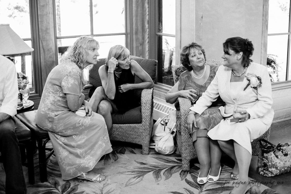 Four women sit and converse in a living room with large windows. Two wear dresses, one in a light floral print and another in black. The third woman is dressed in a suit, and the fourth wears a lace dress. They appear engaged in friendly conversation.