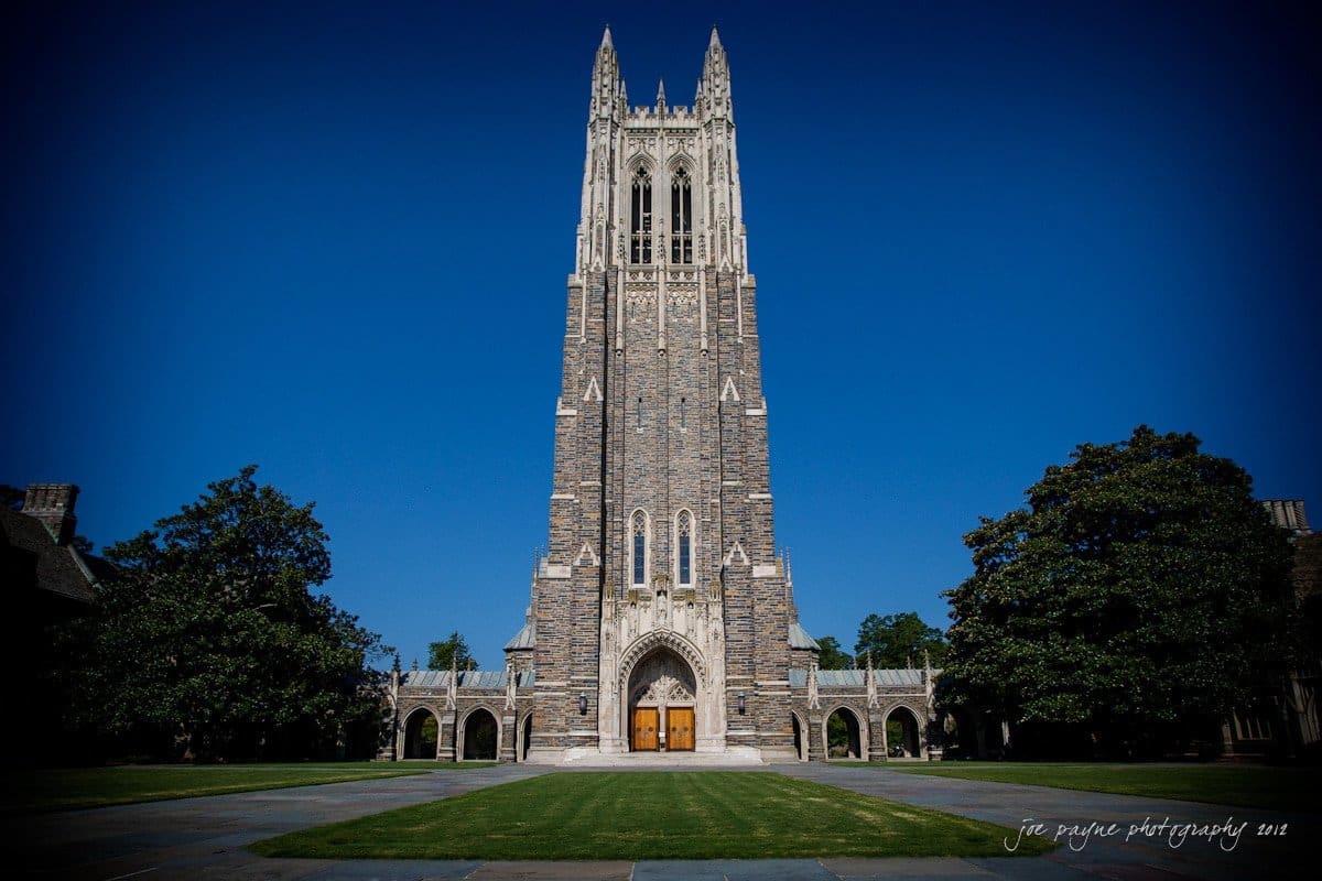 A tall, Gothic-style cathedral with a pointed spire stands against a clear blue sky, flanked by green trees. The entrance is ornate, with large wooden doors, and a paved courtyard leads up to the building.