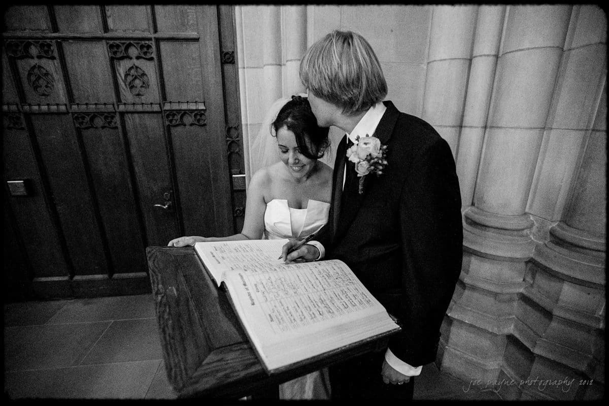 A bride and groom stand next to each other in a church setting, signing a large open book on a wooden stand. The groom points at the page while the bride smiles. They are both dressed in wedding attire.