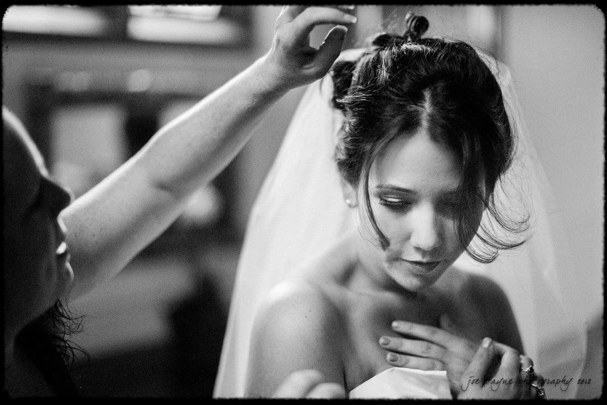 A bride with dark hair is being adjusted by another person, who is styling her hair. She wears a strapless dress and veil, looking down slightly with a serene expression. The black and white image has a soft, intimate feel.