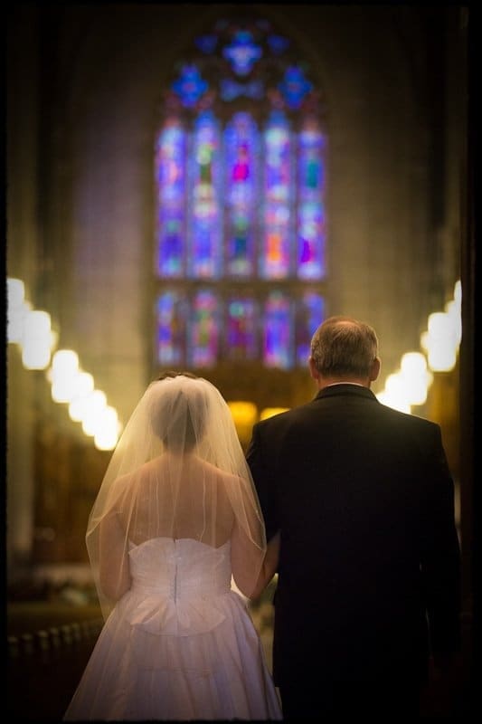 A bride in a white wedding dress and veil walks arm-in-arm with an elderly man down a church aisle. Soft lighting highlights colorful stained glass windows in the background, creating a warm and serene atmosphere.