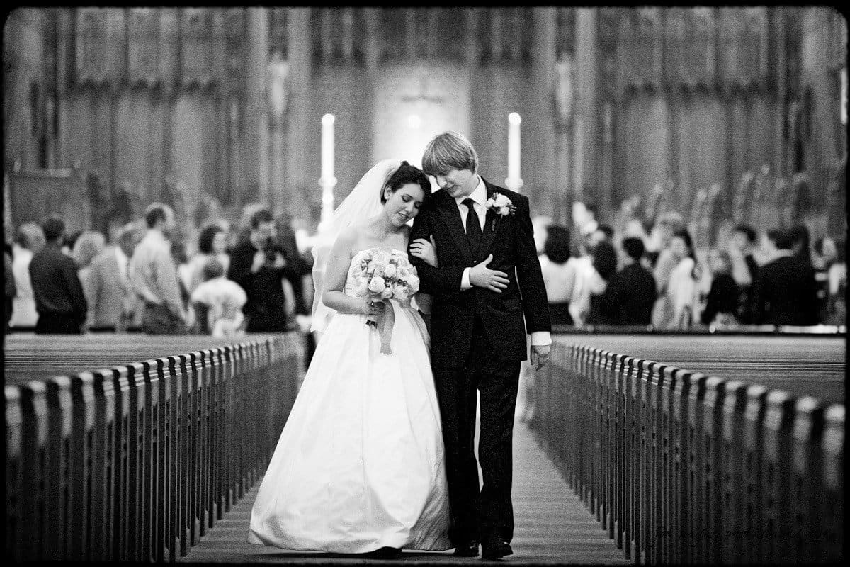 A bride in a white gown and a groom in a dark suit walk arm in arm down the aisle of Duke Chapel, smiling at each other, with wedding guests blurred in the background.
