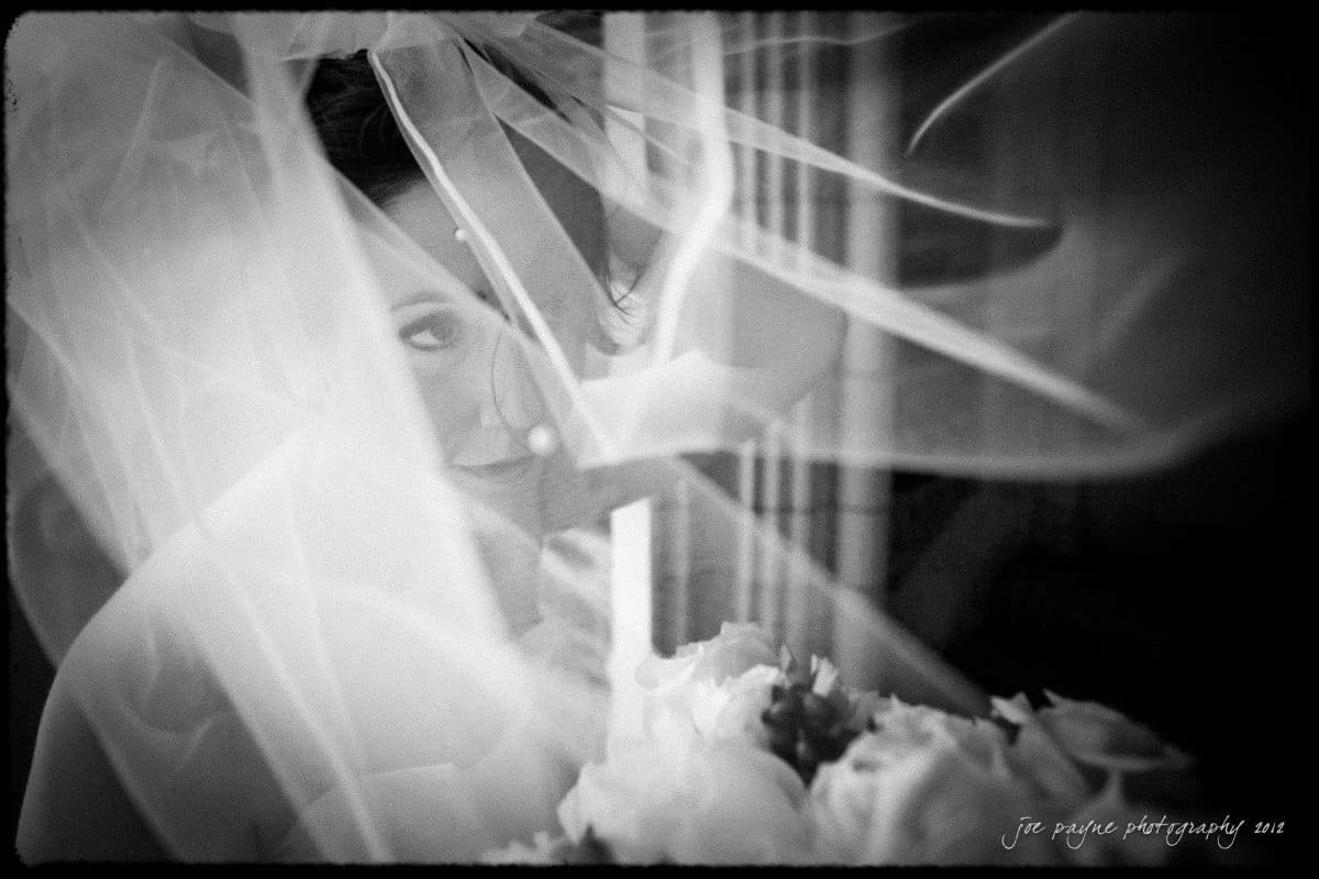 A black and white photo of a bride smiling beneath a flowing veil. The veil softly drapes over her face, partially revealing her eyes and smile. Below the veil, a bouquet of flowers is slightly visible.
